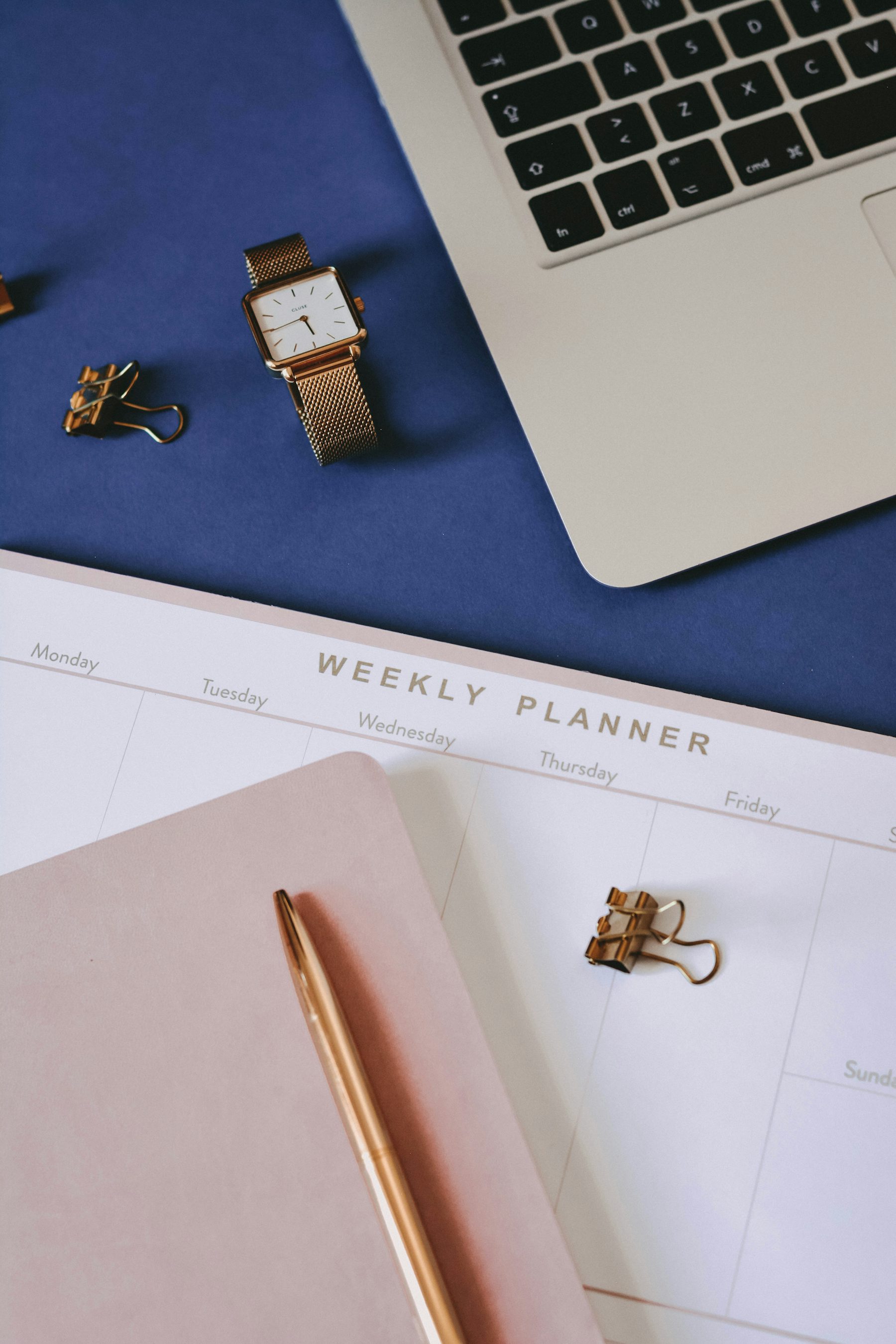 Weekly planner on a desk beside a laptop, a watch and a pen